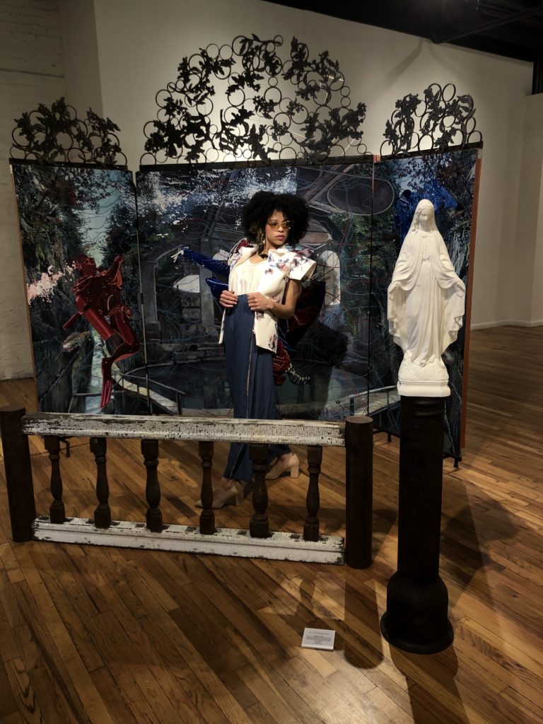 DanShay in a structured neoprene vest and slouchy silk pants, poses in front of art by Melissa Furness